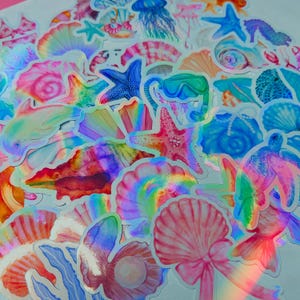 May include: A collection of holographic stickers featuring various sea creatures and shells. The stickers include starfish, seashells, a dolphin, a turtle, and a jellyfish, all in vibrant colors with a shimmering, iridescent effect.