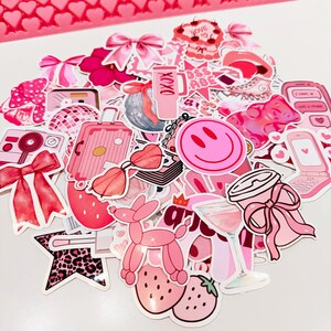 May include: A collection of pink stickers featuring various designs, including a smiley face, a suitcase, a camera, bows, and strawberries. The stickers are in shades of pink and red, with some having glitter accents. The overall theme is girly and fun.