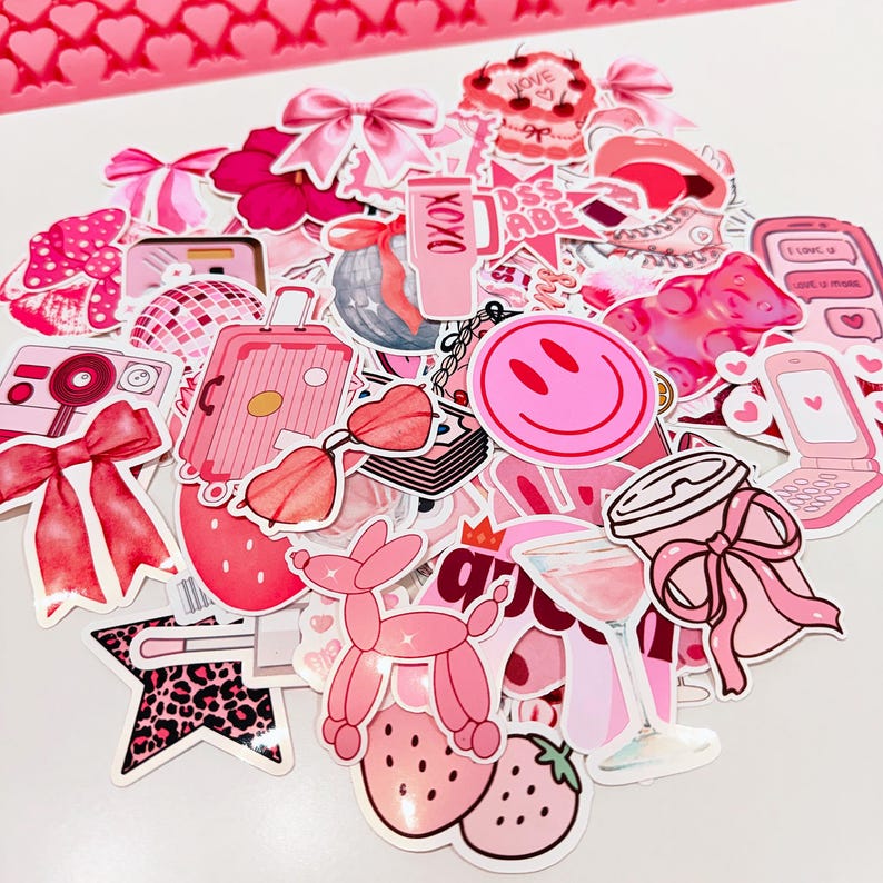 May include: A collection of pink stickers featuring various designs, including a smiley face, a suitcase, a camera, bows, and strawberries. The stickers are in shades of pink and red, with some having glitter accents. The overall theme is girly and fun.