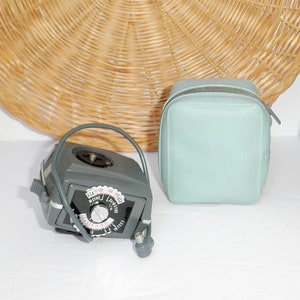 May include: A light blue, rectangular box with a rounded top and a small, gray, round device with a dial and a cord. The box and device are sitting on a white surface. A wicker basket and a gnome figurine are in the background.