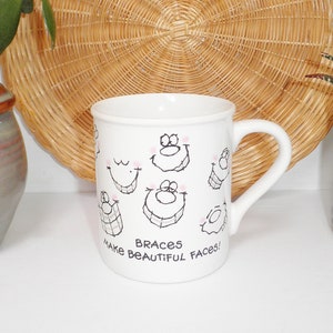 May include: White ceramic mug with a black and white illustration of smiling faces with braces. The text "Braces Make Beautiful Faces!" is printed on the mug.
