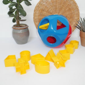 May include: A colorful shape sorter toy with a blue, red, and yellow ball and several yellow geometric shapes. The toy is designed for early childhood development and learning. A woven basket and potted plants are in the background.
