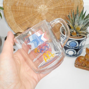 May include: A clear glass mug with a handle. The mug has a colorful graphic of the Olympic rings and a star with red, white, and blue stripes. The text "Games of the XXIIIrd Olympiad Los Angeles 1984" is printed on the mug.