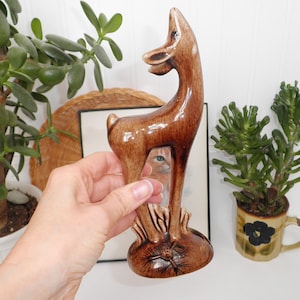 May include: A brown ceramic deer figurine with a glossy finish. The deer has a slender neck and is standing on a textured base. The figurine is held in front of a white wall with plants in the background.