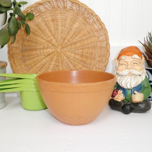 May include: A terracotta colored mixing bowl with a smooth, rounded surface. The bowl is sitting on a white surface with a green plant, a wicker basket, and a gnome figurine in the background.