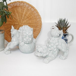 May include: Two white ceramic foo dog figurines. The foo dogs are in a sitting position with their paws raised. One foo dog is holding a ball in its paws. The foo dogs are a light blue color.