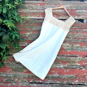 May include: A sleeveless white dress featuring a square neckline with an intricate beige crochet panel across the chest and shoulders. The hem of the dress is finished with a delicate white crochet lace trim, and the garment hangs on a wooden hanger.