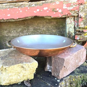 May include: A copper-colored pan with brass handles. The pan has a wide, shallow design and is sitting on a stone surface. The background features weathered wood with peeling red paint. The pan is likely used for cooking.