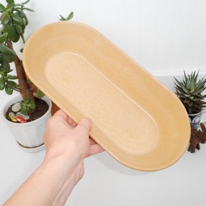 May include: A light brown oval-shaped ceramic serving dish. The dish is smooth and has a slightly textured surface.
