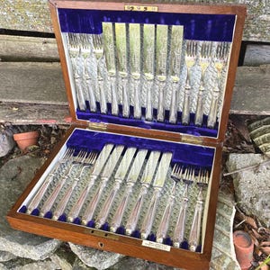 May include: A wooden cutlery case with a velvet interior holds a set of silver forks and knives. The silverware has ornate handles and is arranged neatly within the case. The case is open, revealing the contents.