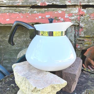 May include: White ceramic coffee pot with a black handle and lid. The pot has a gold band around the middle. The coffee pot is sitting on a light-colored stone and a dark red brick. The background is a weathered wooden wall.
