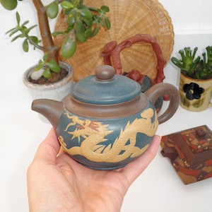 May include: A small, brown and blue teapot with a dragon design. The teapot has a lid and a handle. The dragon is in a light brown color and is on a blue background.