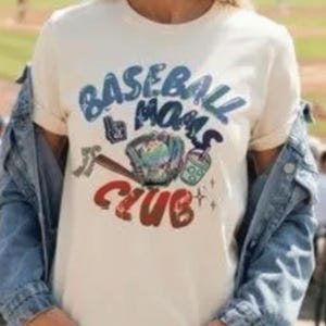 Peut inclure: Un t-shirt à manches courtes couleur crème avec un motif portant l'inscription BASEBALL MOMS CLUB en bleu et rouge, accompagné d'illustrations d'une batte de baseball, d'un gant et d'un gobelet, porté sous une veste en jean bleue.