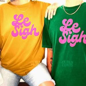 May include: Mustard yellow and green t-shirts with the phrase "Le Sigh" in bright pink, stylized lettering. The green shirt has the text "Curly Coats" below the main text. Casual, unisex apparel.