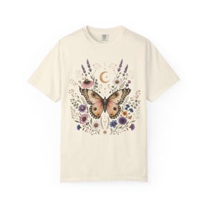 Peut inclure: Un t-shirt à manches courtes de couleur crème arborant un motif central représentant un grand papillon brun entouré de diverses fleurs sauvages colorées, dont de la lavande violette et des marguerites blanches, avec un croissant de lune.