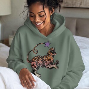 Peut inclure: Un sweat-shirt à capuche vert sauge arborant une impression graphique représentant un léopard au pelage dégradé orange et rose. Le léopard est illustré en plein saut, gueule ouverte, avec une étoile violette située au-dessus de sa queue enroulée.