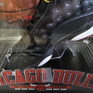 May include: Black and white basketball shoe with red accents. The shoe is in a display case with a red and white Chicago Bulls logo and the text "NBA Finals" and "Champions".