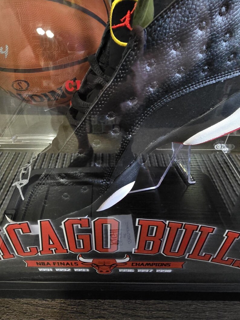 May include: Black and white basketball shoe with red accents. The shoe is in a display case with a red and white Chicago Bulls logo and the text "NBA Finals" and "Champions".