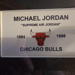 Pu&ograve; includere: Un cartello rettangolare bianco con la scritta "MICHAEL JORDAN" e "SUPREME AIR JORDAN" in nero. Il logo dei Chicago Bulls &egrave; al centro, con gli anni 1984 e 1998 su entrambi i lati. Il cartello ha un bordo argentato.