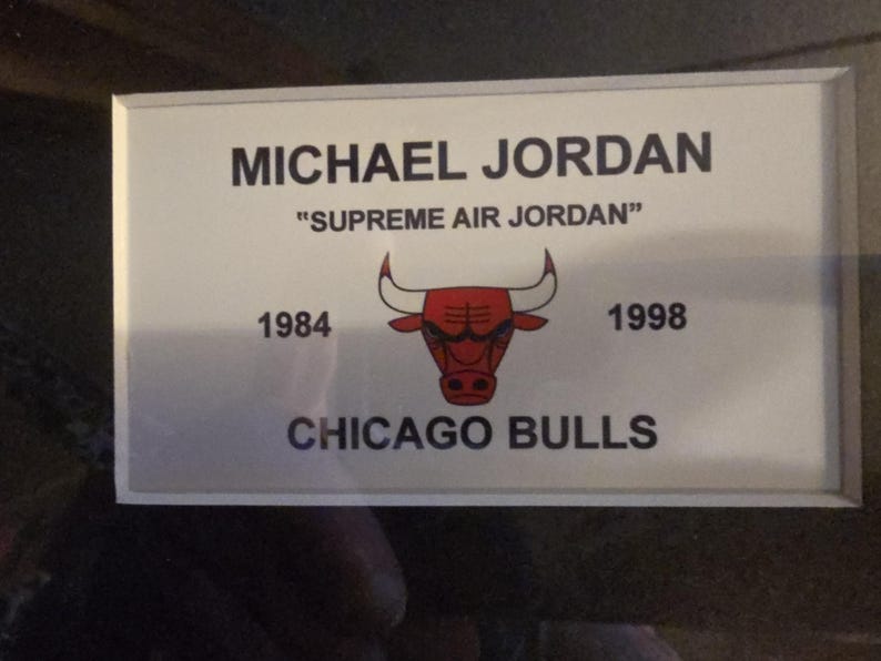 Pu&ograve; includere: Un cartello rettangolare bianco con la scritta "MICHAEL JORDAN" e "SUPREME AIR JORDAN" in nero. Il logo dei Chicago Bulls &egrave; al centro, con gli anni 1984 e 1998 su entrambi i lati. Il cartello ha un bordo argentato.