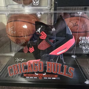 May include: A black and red Air Jordan basketball shoe with a clear display case. The case has a Chicago Bulls logo and the text "NBA Finals Champions 1991 1992 1993 1996 1997 1998".
