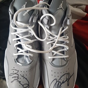 Puede incluir: Un par de zapatillas grises con cordones blancos y una firma negra en cada zapato. La firma en el zapato izquierdo dice "Mike" y la firma en el zapato derecho dice "Jordan".