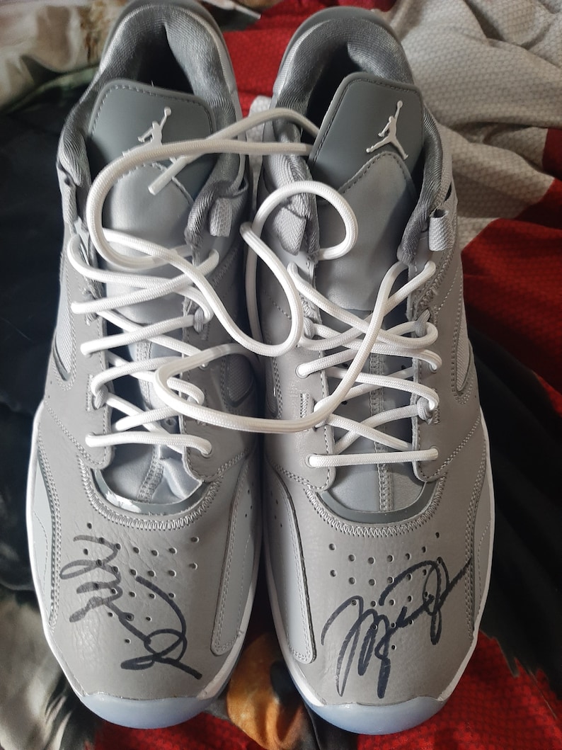 Puede incluir: Un par de zapatillas grises con cordones blancos y una firma negra en cada zapato. La firma en el zapato izquierdo dice "Mike" y la firma en el zapato derecho dice "Jordan".