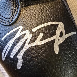 May include: Black leather shoe with a white signature that says "Mike" on the toe. The shoe has a gold "JORDAN" label on the top.