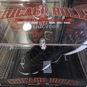 May include: A black and white basketball shoe with a red and white Chicago Bulls logo on the bottom. The shoe is signed in silver marker. The shoe is displayed in a clear acrylic case with a red and white Chicago Bulls logo on the top.