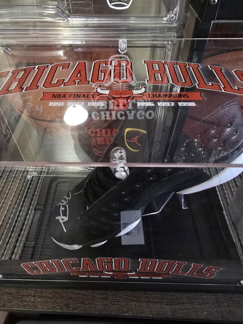 May include: A black and white basketball shoe with a red and white Chicago Bulls logo on the bottom. The shoe is signed in silver marker. The shoe is displayed in a clear acrylic case with a red and white Chicago Bulls logo on the top.