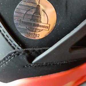 May include: A close-up of a black and red sneaker with a silver sticker that says "Empire Authentics" and the number "101782".