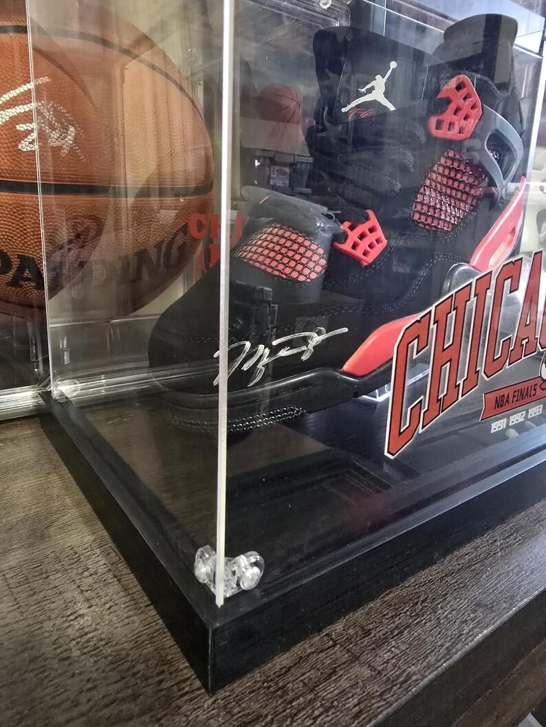 May include: A black and red basketball shoe with a red and white signature on the side. The shoe is in a clear display case with a black base. The shoe is labeled "Chicago" and "NBA Finals 1991".