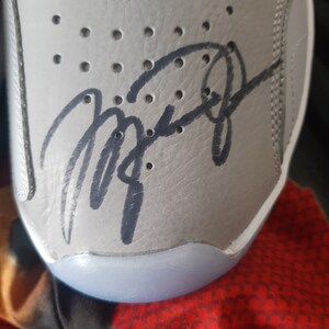 Puede incluir: Primer plano de un zapato de cuero gris con una firma negra en el lateral. La firma est&aacute; escrita en cursiva y dice "Michael Jordan".