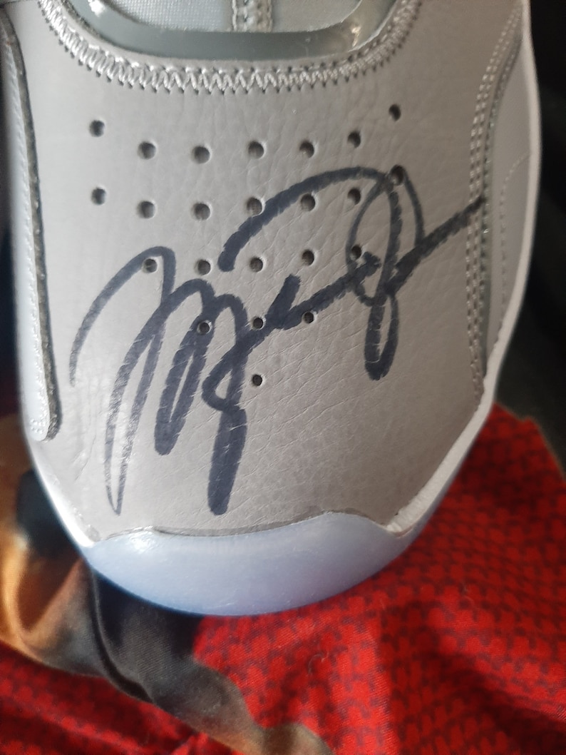 Puede incluir: Primer plano de un zapato de cuero gris con una firma negra en el lateral. La firma est&aacute; escrita en cursiva y dice "Michael Jordan".