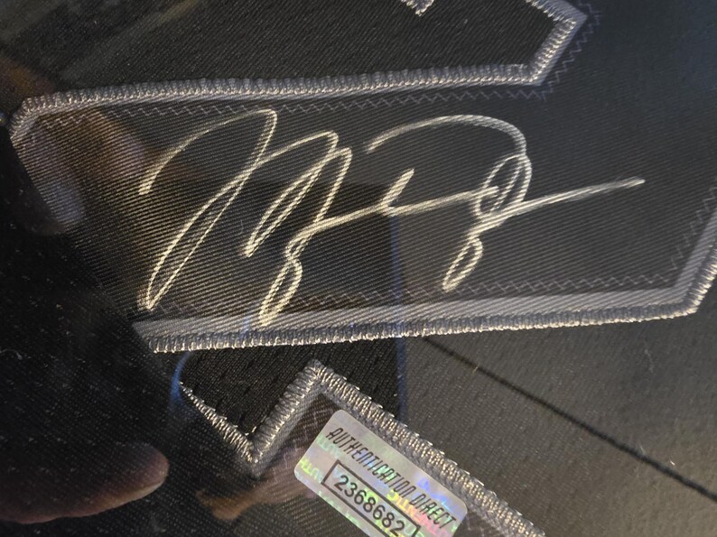 Pu&ograve; includere: Primo piano di un tessuto nero con una firma ricamata bianca. La firma &egrave; su una toppa con un bordo argentato. &Egrave; visibile un adesivo trasparente con la scritta "AUTHENTICATION DIRECT" e un numero di serie.