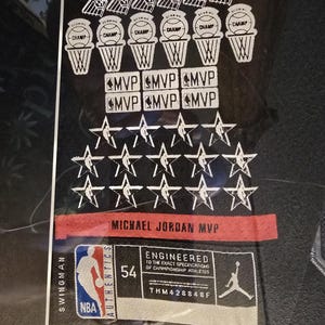 Pu&ograve; includere: Maglia da basket nera con grafica bianca. Presenta canestri da basket, la parola "CHAMP", "MVP" e stelle. Include il testo "MICHAEL JORDAN MVP" e una toppa "54". Sono visibili anche i loghi NBA e Jordan.