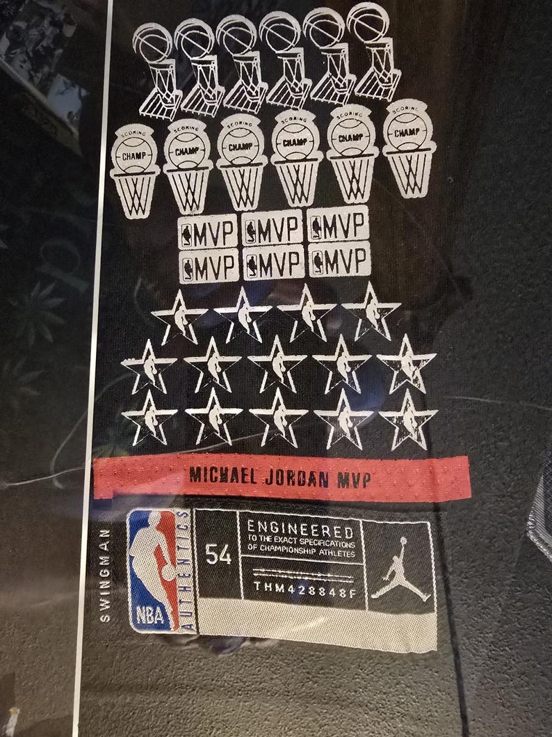 Pu&ograve; includere: Maglia da basket nera con grafica bianca. Presenta canestri da basket, la parola "CHAMP", "MVP" e stelle. Include il testo "MICHAEL JORDAN MVP" e una toppa "54". Sono visibili anche i loghi NBA e Jordan.
