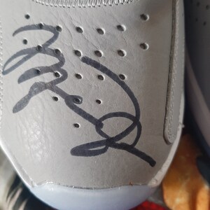 Puede incluir: Primer plano de un zapato de cuero gris con una firma negra en el lateral. El zapato tiene una suela blanca y una fila de peque&ntilde;os agujeros en el lateral.