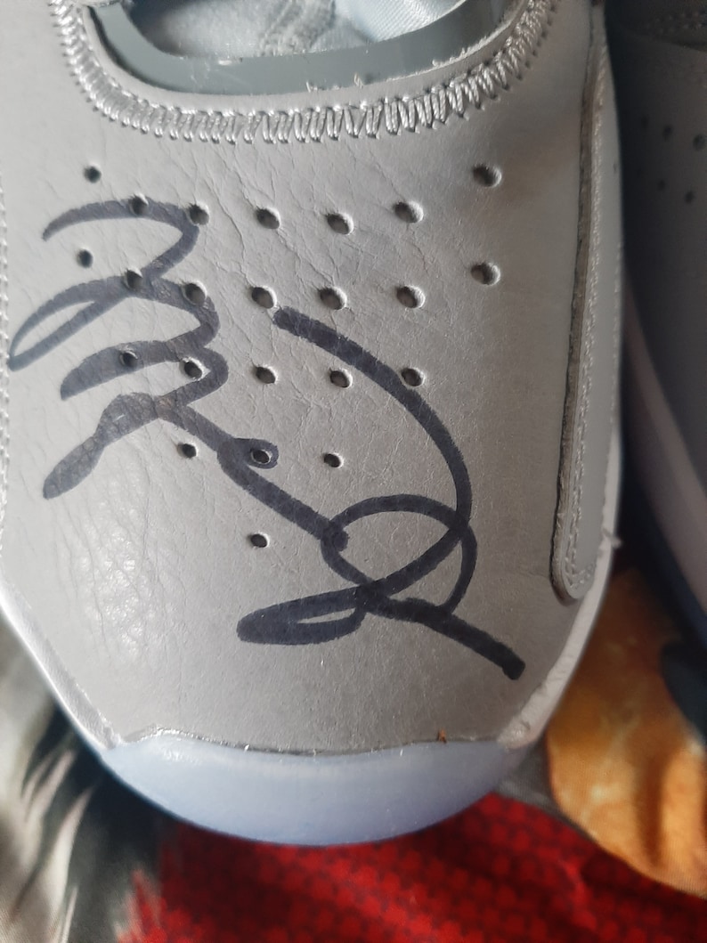 Puede incluir: Primer plano de un zapato de cuero gris con una firma negra en el lateral. El zapato tiene una suela blanca y una fila de peque&ntilde;os agujeros en el lateral.