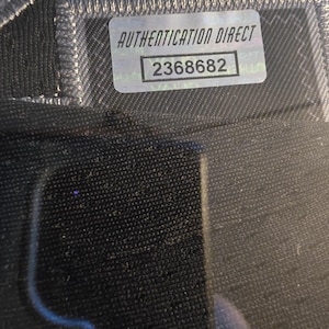 Pu&ograve; includere: Primo piano di un tessuto nero con un adesivo argentato Authentication Direct. L'adesivo riporta la scritta "AUTHENTICATION DIRECT" e il numero "2368682". Il tessuto presenta una cucitura visibile.