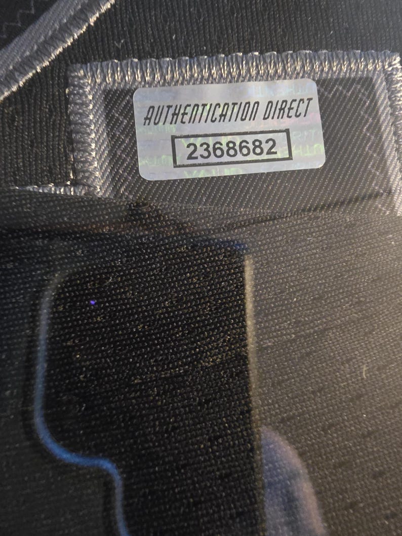 Pu&ograve; includere: Primo piano di un tessuto nero con un adesivo argentato Authentication Direct. L'adesivo riporta la scritta "AUTHENTICATION DIRECT" e il numero "2368682". Il tessuto presenta una cucitura visibile.