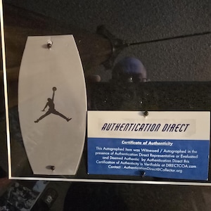 Pu&ograve; includere: Un'esposizione incorniciata con un emblema grigio a forma di scudo con la silhouette nera di una persona che salta con un pallone da basket. Sotto, un certificato di autenticit&agrave; con la scritta "AUTHENTICATION DIRECT".