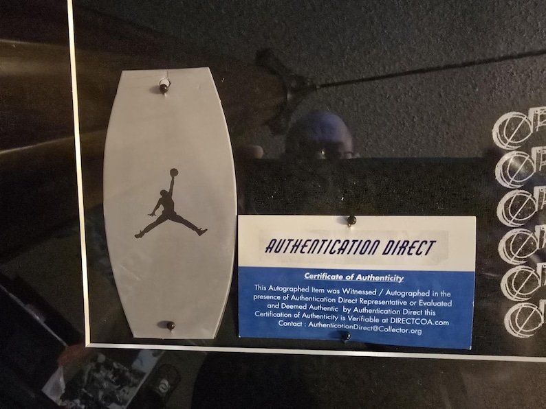Pu&ograve; includere: Un'esposizione incorniciata con un emblema grigio a forma di scudo con la silhouette nera di una persona che salta con un pallone da basket. Sotto, un certificato di autenticit&agrave; con la scritta "AUTHENTICATION DIRECT".