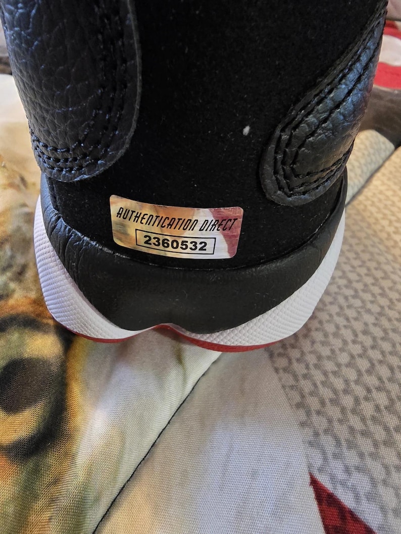 May include: Close-up of the back of a black and white sneaker with a red accent. The sneaker has a tag that reads "AUTHENTICATION DIRECT" and "2360532".
