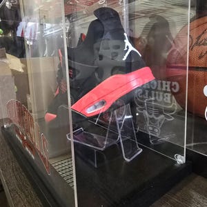 May include: A black and red Air Jordan sneaker with a white logo is displayed in a clear acrylic display case with a black base. The case has a Chicago Bulls logo on the side.