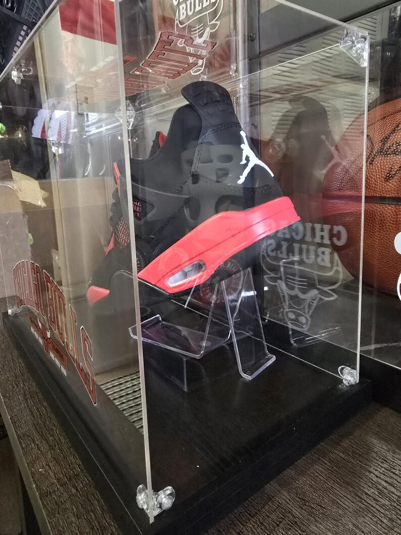May include: A black and red Air Jordan sneaker with a white logo is displayed in a clear acrylic display case with a black base. The case has a Chicago Bulls logo on the side.