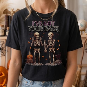 Puede incluir: Una camiseta negra con un gráfico de dos esqueletos, uno sosteniendo la columna vertebral del otro, con el texto I'VE GOT YOUR BACK. El diseño incluye una calabaza sobre un tocón, un gato negro, murciélagos y dulces de maíz dispersos.
