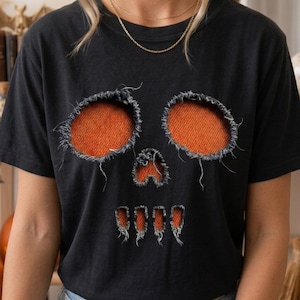 Puede incluir: Una camiseta negra de manga corta con un diseño gráfico de calavera creado con cortes desgastados y deshilachados. Las cuencas de los ojos, la nariz y los dientes revelan una tela naranja brillante y texturizada bajo el material negro.