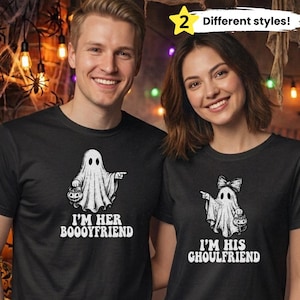 Puede incluir: Dos camisetas negras con gráficos de fantasmas blancos. La camiseta izquierda muestra un fantasma con un cubo de calabaza y el texto I'M HER BOOOYFRIEND. La derecha muestra un fantasma con un lazo y un cubo con el texto I'M HIS GHOULFRIEND.
