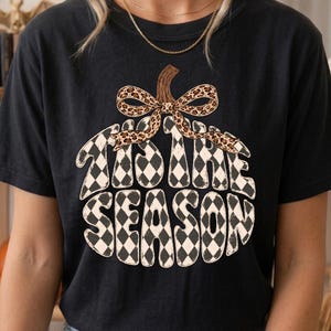 Puede incluir: Una camiseta negra de manga corta con un gráfico en forma de calabaza relleno de un patrón de rombos en blanco y negro. El texto 'TIS THE SEASON' aparece en una fuente negrita dentro de la calabaza, con un tallo marrón y un lazo de estampado leopardo.